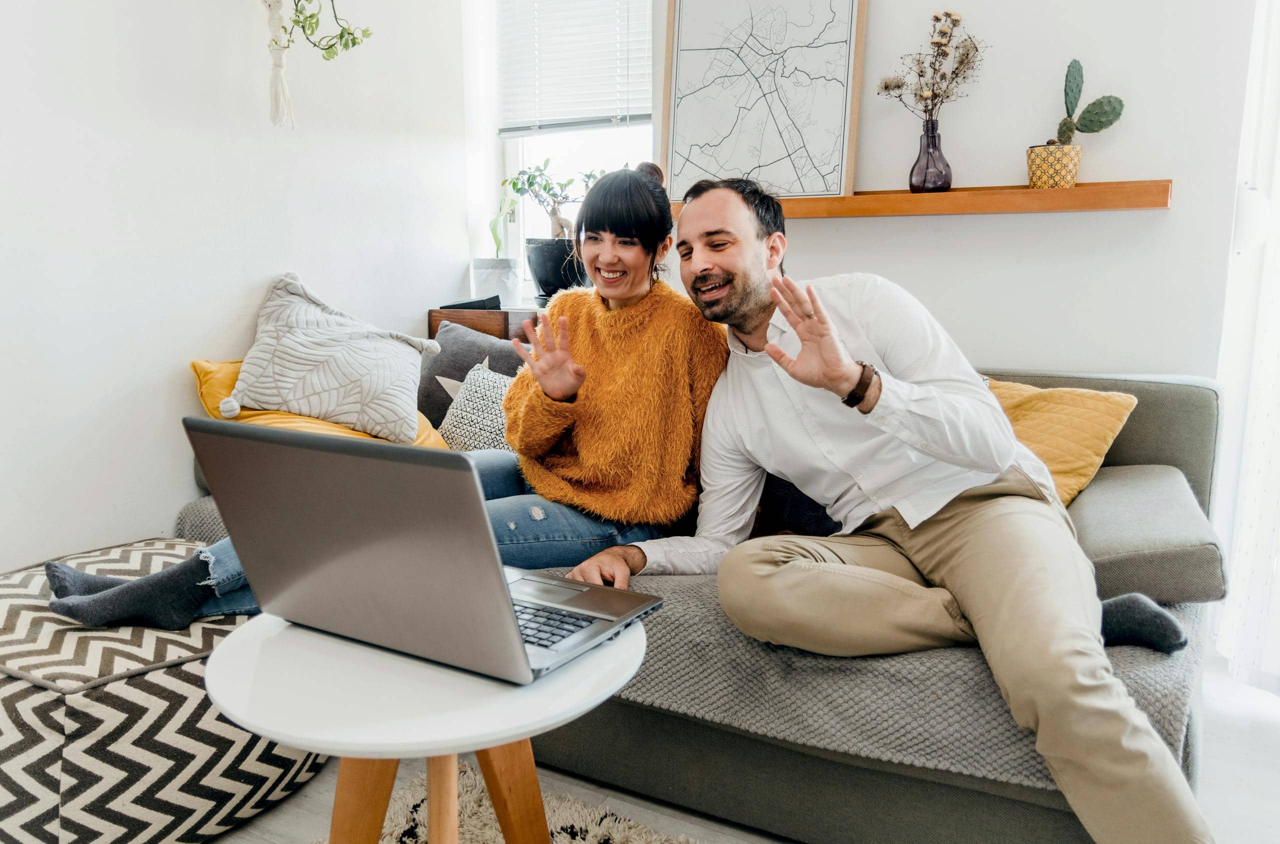 Happy couple sitting on couch, using laptop for a video call in a stylish living room.